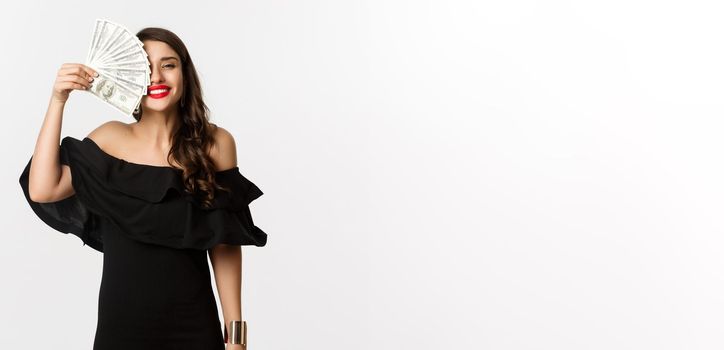 Fashion and shopping concept. Happy young woman in black dress, with red lips, holding money and smiling satisfied, standing over white background.