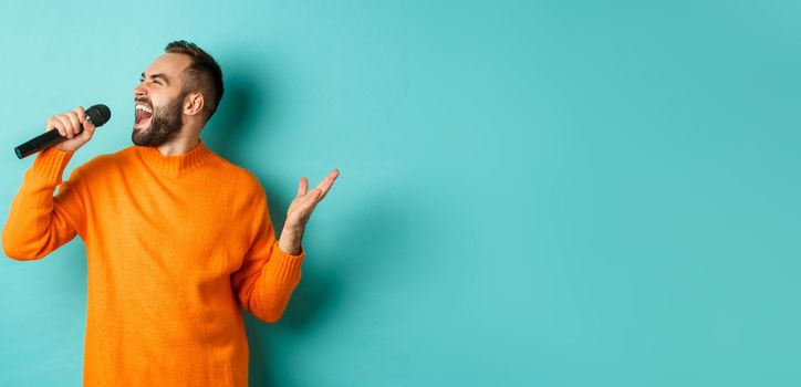 Handsome adult man perform song, singing into microphone, standing against turquoise background. Copy space