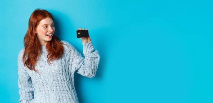 Cute redhead girl in sweater showing credit card, smiling at camera, standing over blue background.