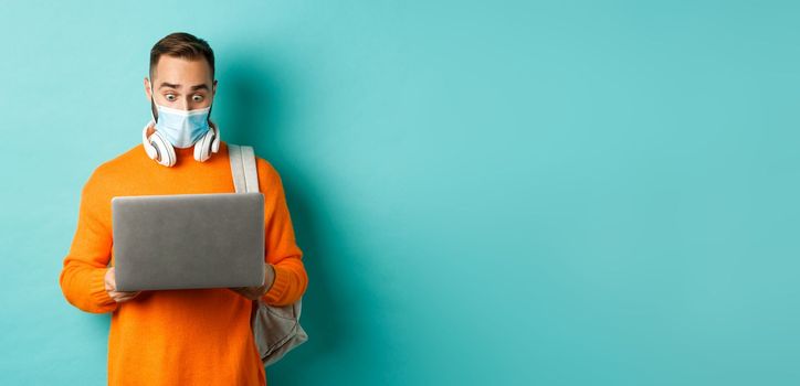 Happy young man in medical mask, working on laptop, looking shocked at computer screen, standing over light blue background.