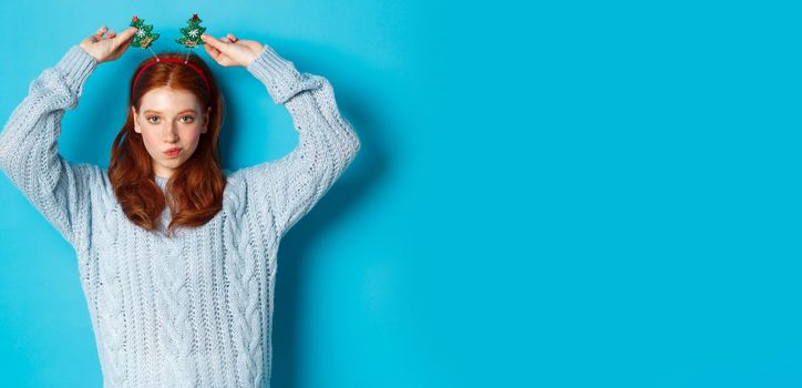 Winter holidays and Christmas sales concept. Beautiful redhead female model celebrating New Year, wearing funny party headband and sweater, smiling at camera.