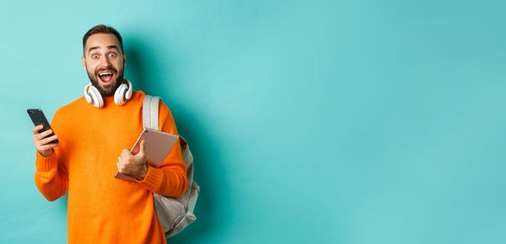 Handsome man student with headphones and backpack, holding digital tablet and smartphone, looking amazed at camera, standing against turquoise background.