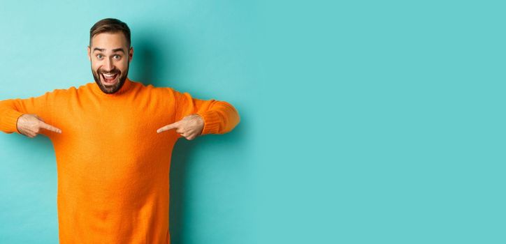 Happy man pointing at sweater, showing your logo banner on clothes, standing over light blue background.