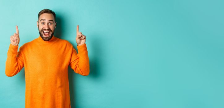 Young happy man showing advertisement, pointing fingers up announcing promo offer, standing over turquoise background.