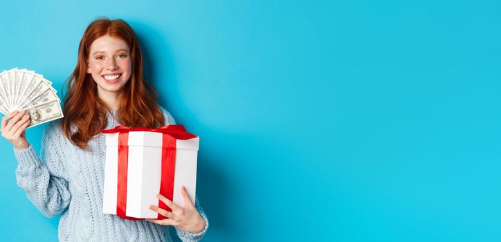 Young redhead woman holding Christmas gift box and money, smiling pleased, standing over blue background.