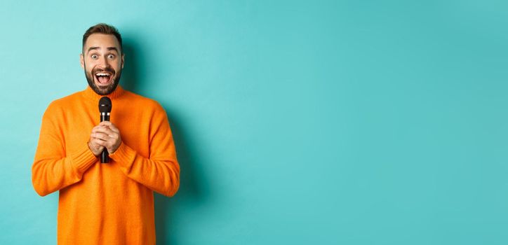 Happy adult man singing karaoke, holding microphone and looking at camera, standing in orange sweater against turquoise background.