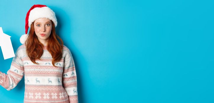 Holiday promos and real estate concept. Cute redhead woman in santa hat and sweater showing paper house model, apartment offer, standing over blue background.