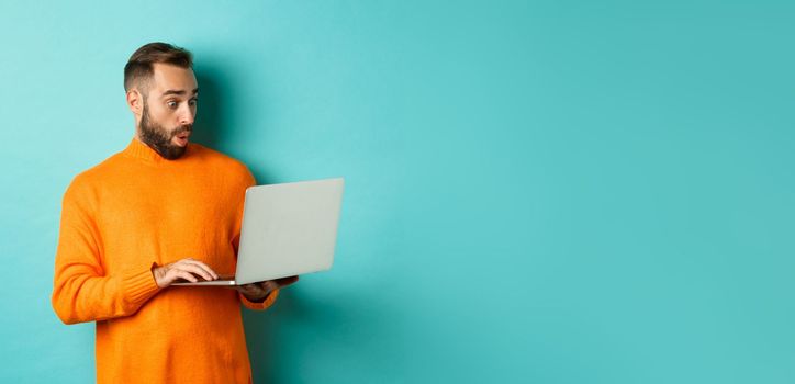 Freelance and technology concept. Image of handsome adult man staring at laptop with amazement, shopping online or working remote, standing over turquoise background.