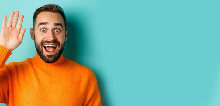 Handsome man waving hand to say hello, giving high five, standing over light blue background.