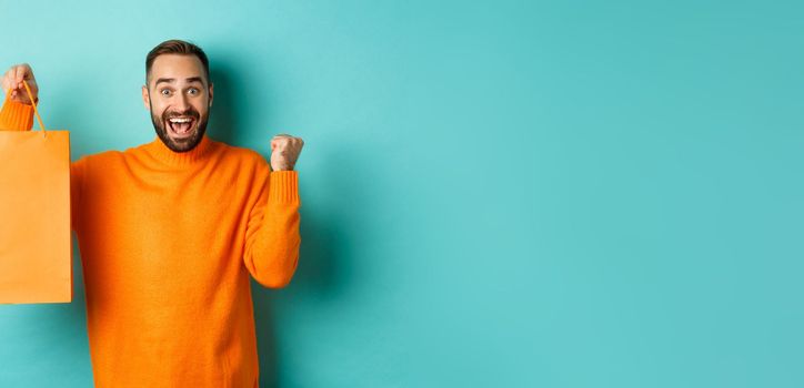 Happy man holding orange shopping bag and rejoicing, got discount and celebrating, standing over turquoise background.