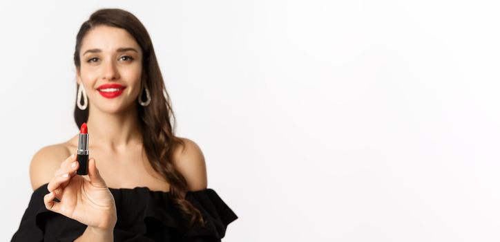 Fashion and beauty concept. Beautiful woman in black dress showing red lipstick and smiling, standing over white background.