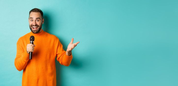 Handsome adult man perform song, singing into microphone, standing against turquoise background.