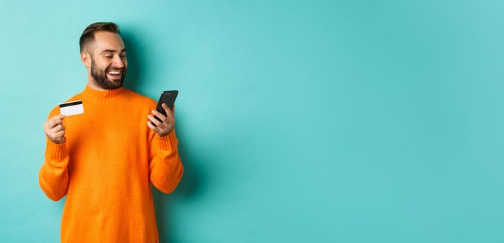 Online shopping. Handsome bearded man paying in internet, holding credit card and stare at mobile screen, standing over turquoise background.