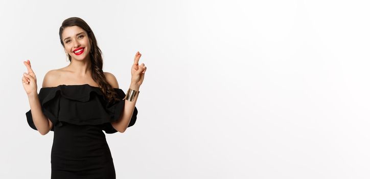 Fashion and beauty. Stylish glamour woman in black dress, red lips, looking optimistic and smiling while cross fingers, making wish, standing over white background.