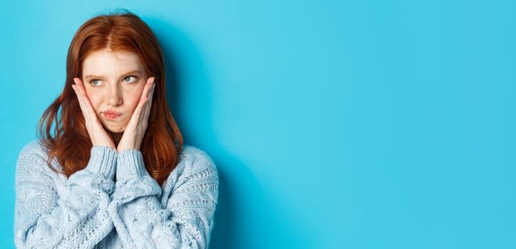 Close-up of troubled teenage girl with red hair staring left, pouting and looking grumpy at promo, standing over blue background.