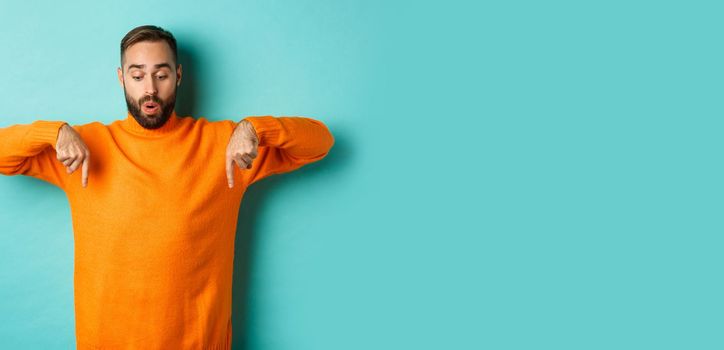 Amazed guy in orange sweater, pointing fingers down and looking with interest at promo, holiday discounts, standing over turquoise background.