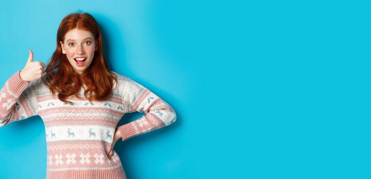 Excited redhead girl showing her support with thumb up, smiling pleased, saying yes in approval, agreeing, standing over blue background.