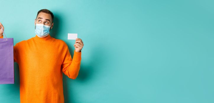 Covid-19, pandemic and lifestyle concept. Thoughtful man in face mask, holding purple shopping bag and credit card, thinking or imaging, standing over turquoise background.