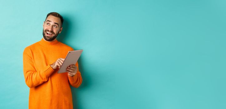 Handsome man thinking and using digital tablet, shopping online and pondering ideas, looking at upper left corner, standing over light blue background.