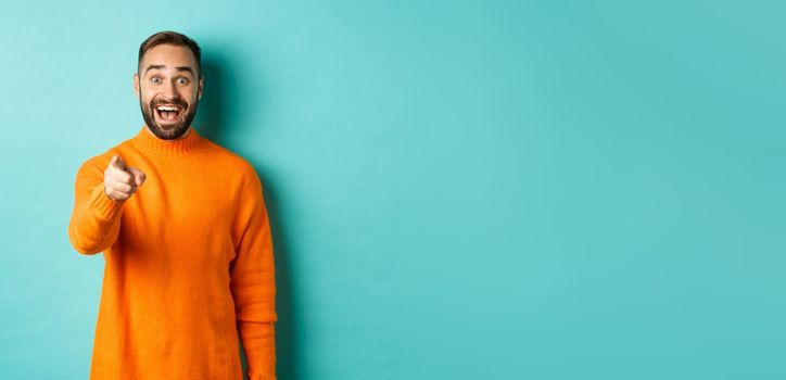 Surprised man stare happy and pointing at camera, recognize someone, excited about seeing something, standing over light blue background.