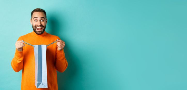 Surprised man open shopping bag and looking amazed, receiving gift on holiday, standing over turquoise background.