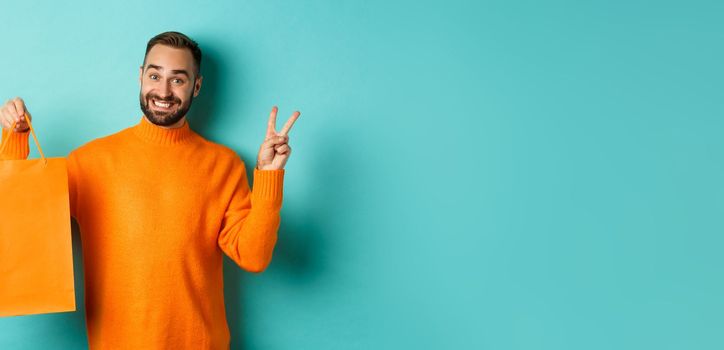 Happy young man showing peace sign and orange shopping bag, smiling pleased, standing over turquoise background. Copy space