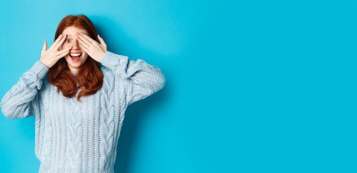 Excited teenage redhead girl open eyes to see holiday surprise, receiving presents, looking amazed at camera, standing over blue background.