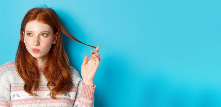 Close-up of thoughtful pretty redhead girl looking left, playing with hair strand and pondering, standing in winter sweater against blue background.