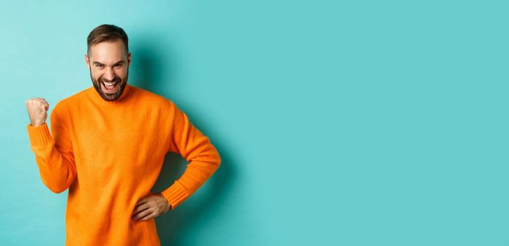 Image of satisfied young man feeling winner, fist pump and saying yes with satisfaction, achieve goal, triumphing while standing over light blue background.