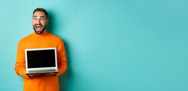 Amazed young man in orange sweater looking at upper left corner, showing laptop screen promo offer, standing over turquoise background.