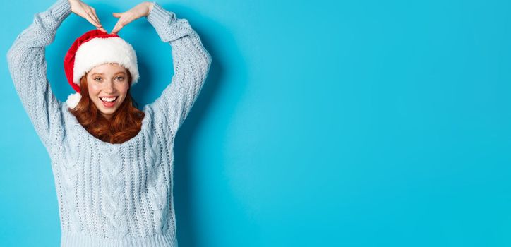 Winter holidays and Christmas Eve concept. Cute redhead teen girl in santa hat and sweater, making heart sign and smiling, wishing merry xmas, standing over blue background.