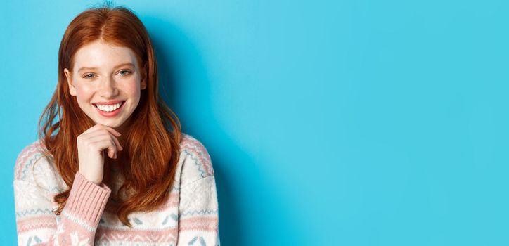 Close-up of beautiful redhead girl smiling, having conversation and staring at camera interested, standing over blue background.