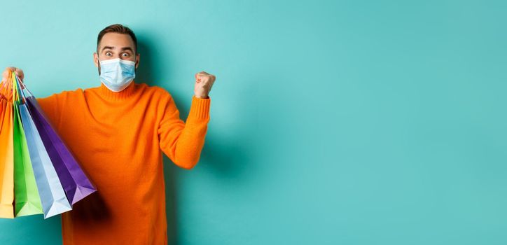 Covid-19, social distancing and lifestyle concept. Excited man in face mask showing shopping bags and rejoicing from discounts, standing over turquoise background.