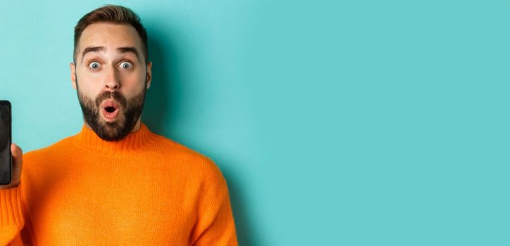 Close-up of handsome bearded guy in orange sweater, showing smartphone screen and smiling, showing promo online, turquoise background.