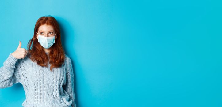 Winter, covid-19 and social distancing concept. Satisfied young redhead woman in face mask showing alright, thumb up gesture and looking left at promo, blue background.