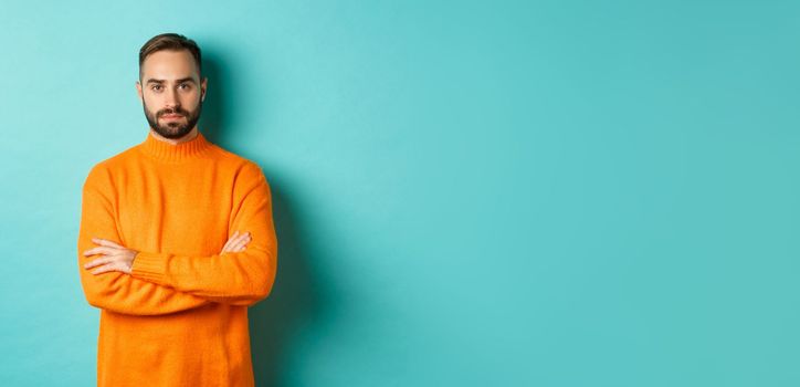 Confident young man looking determined, cross arms on chest, wearing orange winter sweater, standing against turquoise studio background.