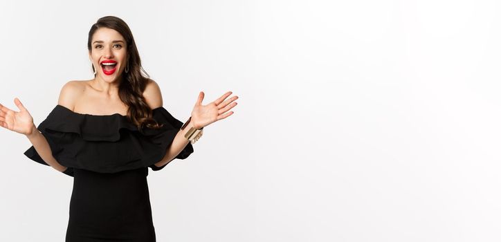 Beauty and fashion concept. Image of surprised and happy young woman in party dress reacting to good news, raising hands up and smiling amazed, white background.