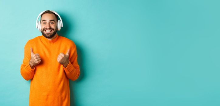Amazed adult man listening music in headphones, looking at camera impressed and showing thumbs-up in approval, recommending, standing over turquoise background.