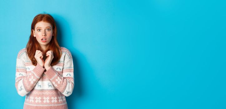 Close-up of cute redhead girl looking with anticipation and worry, staring at camera, standing in winter sweater against blue background.