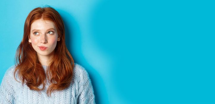 Close-up of cute redhead girl thinking and looking left, have an idea, standing against blue background.