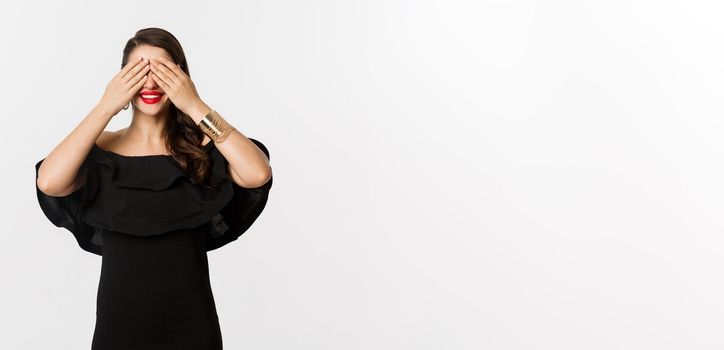 Fashion and beauty. Beautiful woman in black dress and red lipstick, covering eyes and smiling, waiting for surprise, standing over white background.