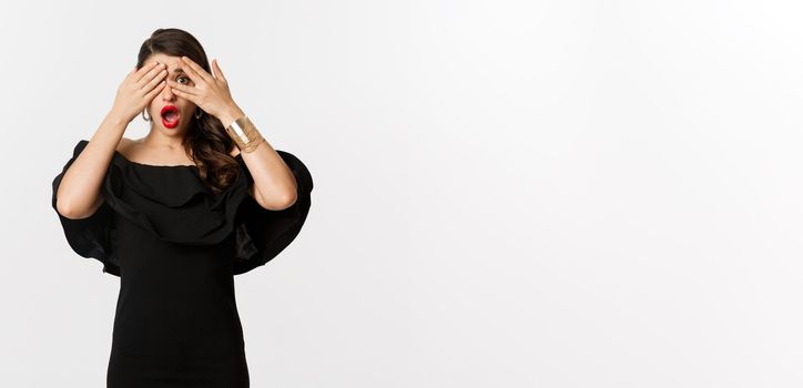 Fashion and beauty. Image of shocked glamour woman in black dress, covering eyes but peeking through fingers startled, standing over white background.
