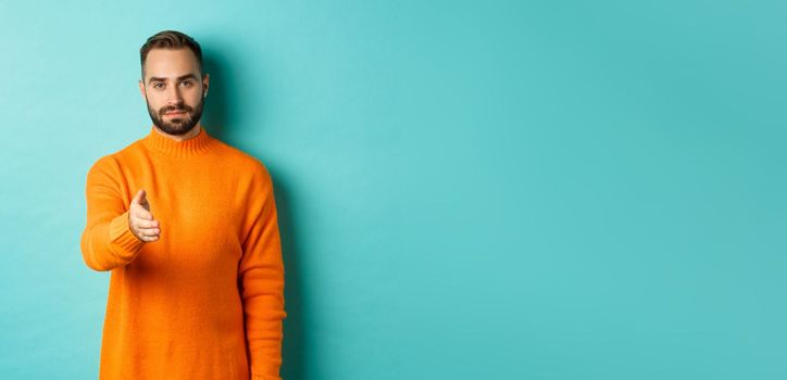 Confident man extending hand for handshake, greeting you, looking self-assured, standing over light blue background. Copy space