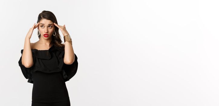 Fashion and beauty. Annoyed and tired woman in black dress, touching head and roll eyes bothered, standing distressed against white background.