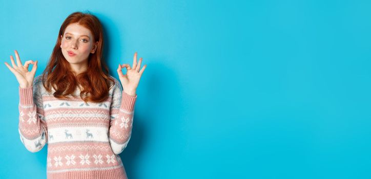 Satisfied and proud redhead girl nod in approval, showing okay sign, not bad or praise gesture, standing against blue background.