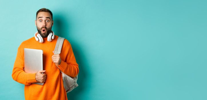 Happy man with backpack and headphones, holding laptop and smiling, looking surprised, standing over turquoise background.