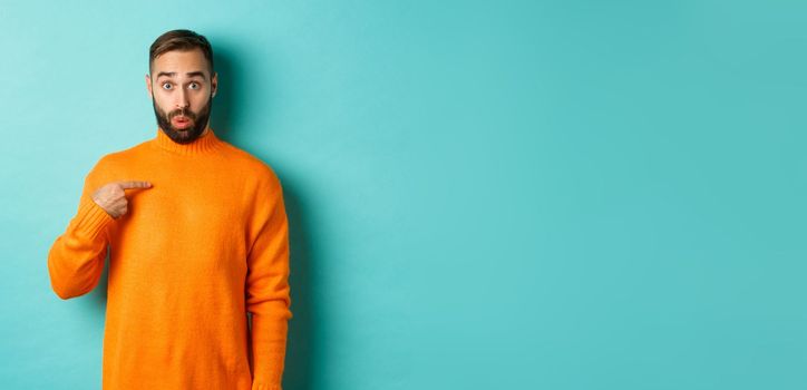 Man pointing at himself with surprise face, being chosen, standing confused against light blue background.