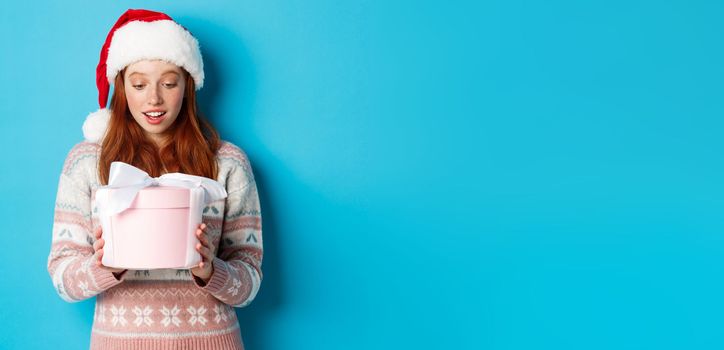 Winter and Christmas Eve concept. Touched and flattered redhead girl looking at box with xmas gift, smiling amazed, standing in santa hat against blue background.