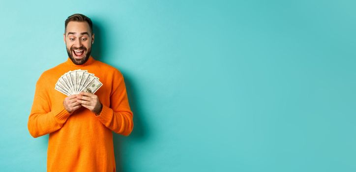 Image of man looking happy at money, smiling amazed, standing over light blue background.