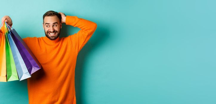 Handsome young man looking awkward and scratching head, staring at shopping bags with gifts, standing over turquoise background.